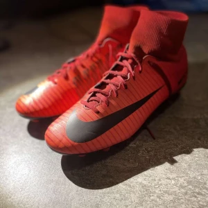 Röda fotbollsskor från Nike - Snygga röda Nike Mercurial fotbollsskor med svart swoosh och snörning. Skorna har en strumpliknande passform och dobbar för optimalt grepp på planen. Perfekta för fotbollsspelare som vill ha stil och funktion.
