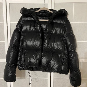Svart pufferjacka från Lawrence Grey - Snygg svart pufferjacka från Lawrence Grey med huva och dragkedja. Jackan har en glansig finish och är perfekt för kyligare väder. Jackan är i storlek M men passar mig som har S perfekt, fungerar nog också om man har XS. Jackan har en perfekt huva som täcker ansiktet när de snöar och regnar så att sminket är skyddat. Jackan är köpt 2023 använd ett få tal gånger och säljs inte längre. 