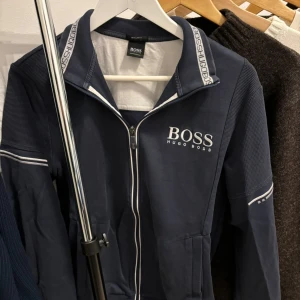Hugo Boss Zip Up tröja - Säljer en stilren mörkblå zipup tröja från Hugo Boss med dragkedja och ribbade detaljer. Tröjan har långa ärmar och märkeslogga på bröstet samt längs kragen. Perfekt för en casual look. I bild 4 & 5 det man minimalistisk slitage pga tvätt (som inte synd för ögat). Nypris: 1499kr
