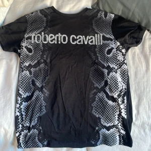Svart t-shirt med ormmönster från Roberto Cavalli - Snygg svart t-shirt från Roberto Cavalli med ett coolt ormmönster i grått och vitt. T-shirten har korta ärmar och ett stort Roberto Cavalli-tryck på framsidan. Perfekt för en trendig look! Andvänt den en gång nytt skick