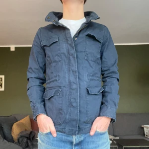 Mörkblå Fieldjacka från Dobber - Säljer en stilren mörkblå Fieldjacka från Dobber i storlek XS. Jackan har en klassisk design med flera fickor och en hög krage. Perfekt för våren som kommer. Hör av er vid frågor!👍