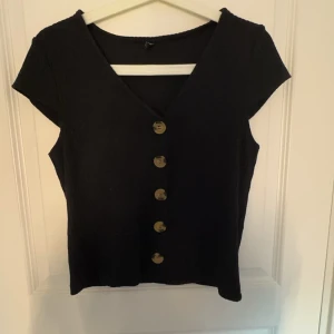 Svart kortärmad topp med knappar - Snygg svart topp med korta ärmar och v-ringning. Framsidan pryds av stora knappar som ger en stilren look. Perfekt för både vardag och festliga tillfällen.