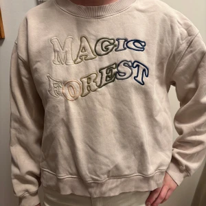 Ljusrosa/beige tröja med brodyr - Säljer en sweatshirt med broderad text 'Magic Forest' i olika färger. Tröjan har långa ärmar och en rund halsringning. Perfekt för en avslappnad stil. Från coaster Copenhagen