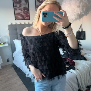 Svart offshoulder topp med fransar - Trendig svart offshoulder topp med fransar som ger en unik look. Perfekt för att sticka ut på festen. Toppen har en lös passform och är tillverkad i ett lätt material.