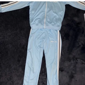 Ljusblå träningsjacka från Palm Angels - Snygg ljusblå träningsjacka från Palm Angels med dragkedja och vita detaljer. Perfekt för en sportig look. Matchande byxor finns också. Det är en stolek M på båda 