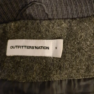 Grå jacka från Outfitters Nation - Snygg grå jacka från Outfitters Nation med knappar och bälte i midjan. Jackan har långa ärmar och en hög krage med ribbade detaljer. Perfekt för kyligare dagar.
