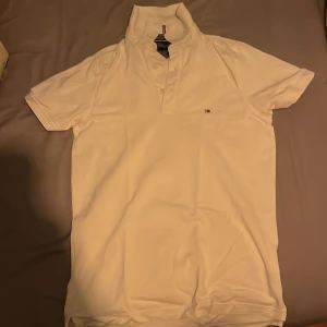 Vit kortärmad pikétröja från Tommy Hilfiger - Snygg vit pikétröja från Tommy Hilfiger med korta ärmar och klassisk krage. Tröjan har en liten broderad logotyp på bröstet och knappar vid halsen. Perfekt för en stilren look.