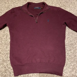 Vinröd tröja från Polo Ralph Lauren - Snygg vinröd tröja från Polo Ralph Lauren med dragkedja vid halsen och broderad logga på bröstet. Perfekt för kyligare dagar med sin stilrena design och bekväma passform.
