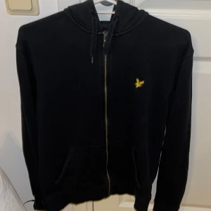Svart hoodie från Lyle & Scott - Säljer en stilren svart hoodie från Lyle & Scott med dragkedja och gul broderad logga på bröstet. Perfekt för en avslappnad look. Pris går att diskuteras 