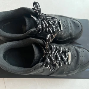 Nike Air Force 1 Low Supreme Black - Säljer ett par svarta Nike Air Force 1 Low Supreme sneakers. Skorna har snygga Supreme-detaljer på snörena och är tillverkade i skinn. Perfekta för dig som gillar stilrena och exklusiva sneakers.