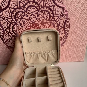 Beige smyckeskrin med dragkedja - Säljer ett superpraktiskt och stilrent beige smyckeskrin med dragkedja. Perfekt för att organisera dina smycken med flera fack och hållare för ringar och örhängen. Skrinet är kompakt och lätt att ta med sig på resor. Bakgrunden har ett snyggt rosa mönster som ger en fin touch.