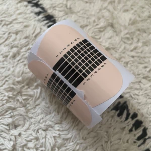 Självhäftande nagelformsmallar - Rulle med självhäftande nagelformsmallar i beige och svart. Perfekt för att skapa jämna och snygga nagelförlängningar. Mallarna har tydliga markeringar för exakt applicering och är enkla att använda. Perfekt för både nybörjare och proffs!