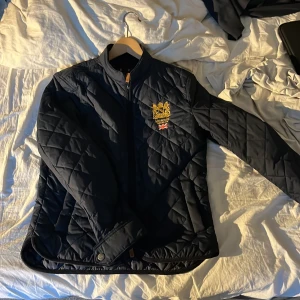 Svart quiltad jacka från Morris - Säljer en snygg svart quiltad jacka från Morris. Den har ett broderat emblem på bröstet och en dragkedja framtill. Jackan är perfekt för höst och vår, med långa ärmar och en stilren design. Passar både till vardags och fest!