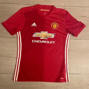 Röd Manchester United fotbollströja - Säljer en snygg röd Manchester United fotbollströja från Adidas. Den har korta ärmar och är i bra skick. Tröjan har det klassiska klubbmärket på bröstet. Lite slitet vid trycket. Perfekt för alla United-fans!