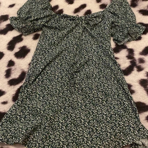 Grön klänning  - Fin grön klänning med vita blommor, knäp och dragkedja, och ett smöre, fint och rent skick, inte trasig, använd några få tal gånger. Pris går att diskutera. 