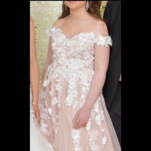 Champagnefärgad balklänning med blommig spets - Elegant champagnefärgad balklänning med offshoulder-design och blommig spetsdetalj. Klänningen har en lång, flödande kjol som ger en romantisk känsla. Perfekt för speciella tillfällen.