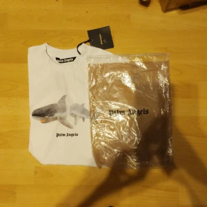 Vit t-shirt med hajmotiv från Palm Angels - Säljer en cool vit t-shirt från Palm Angels med ett unikt hajmotiv på framsidan. Pris kan diskuteras vid snabbt köp.                 T-shirten har en klassisk passform och är kortärmad, perfekt för en avslappnad stil.