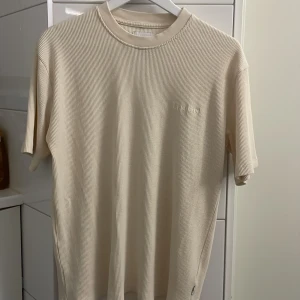 Beige ribbad t-shirt från Woodbird - Säljer en stilren beige ribbad t-shirt från Woodbird. Den har en klassisk rund hals och korta ärmar. Perfekt för en avslappnad look. Passar bra till jeans eller shorts. Storlek XS men passar S och M.