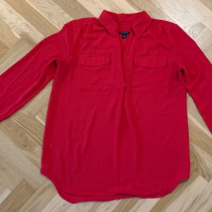 Röd blus från Ann Taylor - Röd blus från Ann Taylor med långa ärmar och klassisk krage. Blusen har två bröstfickor och en avslappnad passform. Material: Polyester