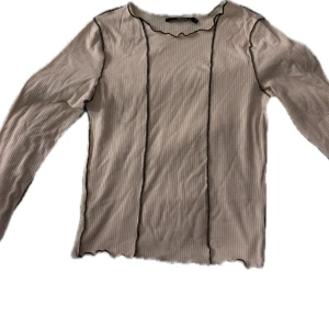 Beige ribbad tröja med mörkbruna detaljer - Säljer en beige ribbad tröja med bruna kontrastsömmar. Tröjan har långa ärmar och en vågig kant vid halsen och ärmsluten. Perfekt för en stilren look!