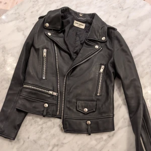 Svart skinnjacka från Saint Laurent - Snygg svart skinnjacka från Saint Laurent med coola dragkedjor och knappar. Jackan har en klassisk bikerstil med asymmetrisk dragkedja och flera fickor. Perfekt för att ge din outfit en edgy touch.