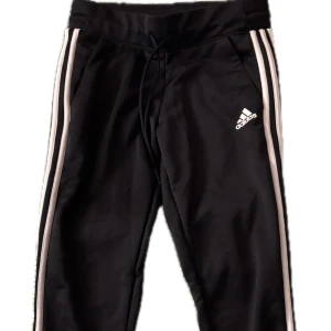 Svarta träningsbyxor från Adidas - Snygga svarta träningsbyxor från Adidas med vita ränder längs sidorna och logga på benet. Byxorna har en bekväm passform med dragsko i midjan för justerbarhet. Perfekta för sport och fritid.