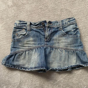 Jeanskjol från Only - Snygg jeanskjol från Only i blå denim med volangdetalj. Kjolen har en klassisk femficksdesign och stängs med knapp och dragkedja framtill. Perfekt för en avslappnad stil. Längd 30 midjemått 32