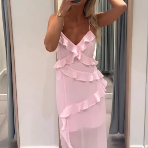 Ljusrosa långklänning med volanger - Elegant ljusrosa långklänning med smala axelband och volangdetaljer framtill. Klänningen har en öppen rygg med knytband, vilket ger en romantisk känsla. Perfekt för sommarens festligheter. Vero Moda.