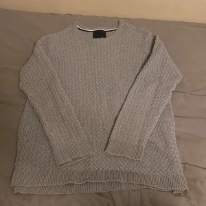 Grå stickad tröja  - Säljer en stilren grå stickad tröja från 157. Tröjan har ett diskret mönster och ribbade kanter vid ärmslut och nederkant. Perfekt för kyligare dagar. Storlek L