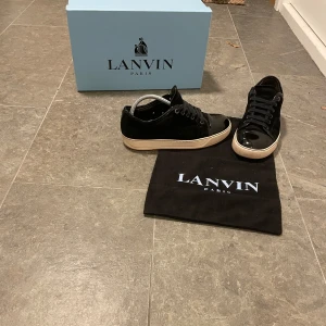 Svarta sneakers från Lanvin - Snygga svarta sneakers från Lanvin med glansig toebox och klassisk snörning. Skorna har en vit sula som kontrasterar snyggt mot det svarta. Perfekta för en stilren look. Storlek 40. Box,dustbag medföljer. Om du har minsta lilla fundering e de bara att skriva.