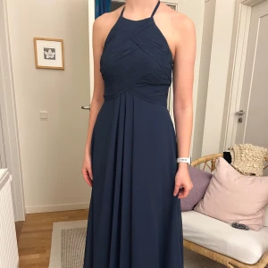 Mörkblå långklänning - Elegant mörkblå långklänning med halterneck och öppen rygg. Perfekt för bal eller fin sittning. Klänningen har en vacker drapering framtill och en dragkedja i ryggen för enkel påklädning. Använd 1 gång. 