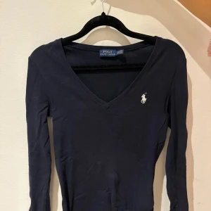 Mörkblå långärmad topp från Ralph Lauren - Säljer en stilren mörkblå långärmad topp från Ralph Lauren med v-ringning och det klassiska logotypbroderiet på bröstet. Perfekt för en elegant och avslappnad look.
