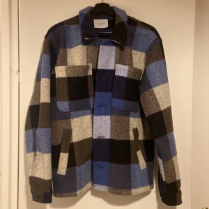 Rutig overshirt från Carlings - Snygg rutig overshirt i blå, svart och grå från Carlings. Jackan har långa ärmar och knappar framtill. Tillverkad i en blandning av återvunnen polyester och ull, vilket ger en skön och hållbar känsla. Perfekt för lager-på-lager under kyligare dagar.