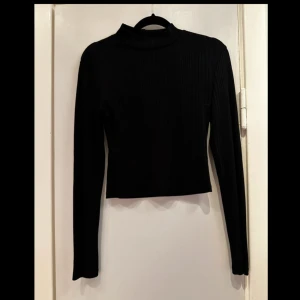 Svart ribbad topp från H&M Divided - Säljer en stilren svart ribbad topp från H&M Divided. Tröjan har en hög krage och långa ärmar, perfekt för en minimalistisk look. Materialet är en blandning av polyester och viskos vilket ger en skön passform.  Mått:  Byst: 38 Midja: 35 cm Längd: 43 cm Innerärm: 47 cm