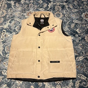 Beige dunväst från Canada Goose - Säljer en snygg beige dunväst från Canada Goose med knappar och broderad logga på bröstet. Västen har två stora fickor framtill och en hög krage för extra värme. Perfekt för kyliga dagar!        Den är lite smutsig men det är bara o lägga den i tvätten så försvinner det dens färg ser inte ut så i verkligheten endast pågrund av ljuset     Jag är rätt så säker på att den är äkta men eftersom jag inte ör helt 100 så är priset lågt 