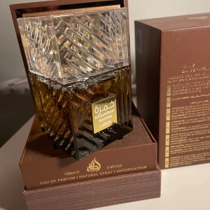 Lattafa Khamrah Qahwa - Elegant och lyxig parfymflaska med ett intrikat mönster och en klar, fyrkantig kork. Flaskan är i en djup bärnstensfärg och rymmer 100 ml. Förpackningen är stilren i brunt med gyllene detaljer. Perfekt för den som söker en sofistikerad doftupplevelse. 98ml kvar! Dma för mer info