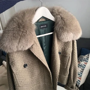 Beige kappa med pälskrage från MEOTINE - Säljer en elegant beige kappa från MEOTINE med en lyxig pälskrage. Jackan har en dubbelknäppning framtill och stora fickor. Materialet är en blandning av ull och syntet, vilket ger en skön och varm känsla. Perfekt för kyliga dagar!