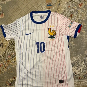 Vit och randig fotbollströja med nummer 10 - Snygg vit fotbollströja från Nike med blå och röda ränder. Tröjan har korta ärmar med blå och röda detaljer. Den pryds av en tupp och nummer 10 på framsidan samt namnet 'MBAPPE' och nummer 10 på baksidan. Perfekt för fotbollsfans!