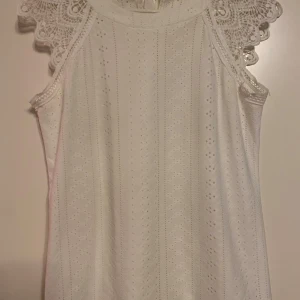 Vit ärmlös topp med spetsdetaljer - Säljer en söt vit ärmlös topp med vackra spetsdetaljer vid axlarna. Toppen har en knytning i nacken. Strl S och fint skick! 