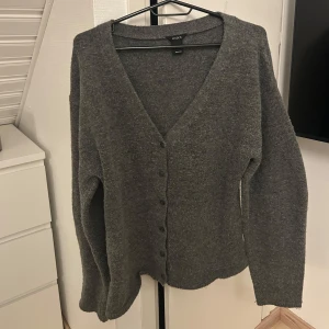 Grå kofta från Lindex - Mysig grå kofta från Lindex med knappar framtill. Perfekt för kyliga dagar med sin mjuka och bekväma passform. Den har en v-ringad design och långa ärmar.