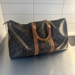 Brun monogramväska från Louis Vuitton - Säljer en klassisk brun monogramväska från Louis Vuitton med läderdetaljer. Väskan har två handtag och en dragkedja för säker förvaring. Dragkedjan är dock lite trasig (se bild) går enkelt att lämna in och fixa har bara inte orkat då den används som prydnad