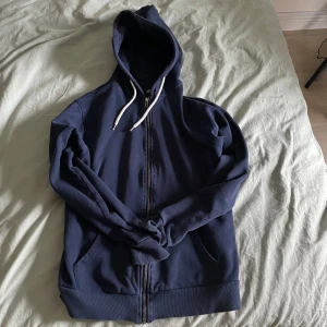 Mörkblå hoodie med dragkedja - Säljer en mörkblå hoodie från lager 157 med dragkedja och huva. Tröjan har långa ärmar och ribbade muddar vid ärmslut och nederkant. Perfekt för en avslappnad stil. Passar på alla stilar och är väldigt clean. 