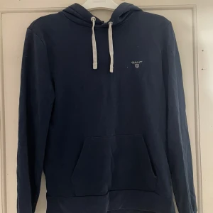 Mörkblå hoodie från GANT - Säljer en snygg mörkblå hoodie från GANT med vit logga på bröstet. Tröjan har en klassisk känguruficka och justerbar huva med vita snören. Perfekt för en avslappnad stil.