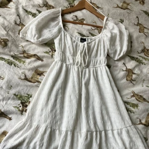 Vit klänning från Gina Tricot - Säljer en söt vit klänning från Gina Tricot i storlek XS. Klänningen har puffiga kortärmar och en knytning framtill. Perfekt för sommarens varma dagar med sin luftiga design och mjuka material.