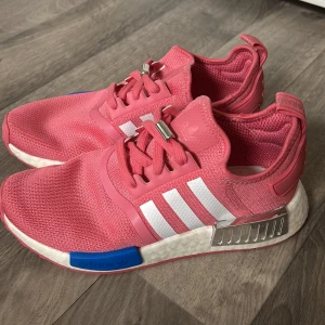 Rosa Adidas NMD sneakers - Snygga rosa Adidas NMD sneakers med vita detaljer och en blå accent på sulan. Skorna har en modern design med bekväm passform och snörning. Perfekta för en sportig look.