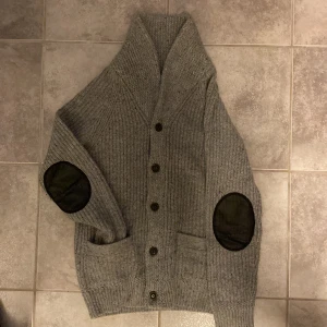 Stickad cardigan - Säljer en stilren grå stickad kofta. Koftan har en mysig sjalkrage och knappar framtill. Den är utrustad med praktiska fickor och har svarta armbågslappar som en snygg detalj. Perfekt för kyliga dagar! För mer frågor eller funderingar hör gärna av dig! MVH Viktor🍀