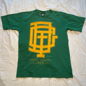 Grön t-shirt med gult tryck från DIL - Säljer en grön t-shirt från DIL med ett stort gult grafiskt tryck på framsidan. T-shirten är kortärmad och tillverkad av ett hampabaserat material. Perfekt för en avslappnad streetwear-look.