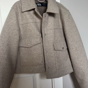Beige jacka från Zara - Snygg beige jacka från Zara med knappar framtill och två fickor med lock. Jackan har en klassisk krage och är perfekt för höst och vår. Den är tillverkad i ett mjukt material som ger en stilren look.