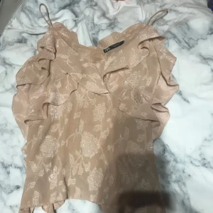 Beige blommig topp från Zara - Säljer en söt beige topp från Zara med blommigt mönster och volangdetaljer. Toppen har smala axelband och en romantisk stil. Perfekt för sommarens alla tillfällen!