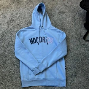 Ljusblå hoodie från Hoodrich - Säljer en snygg ljusblå hoodie från Hoodrich med texten 'HOODRICH'på bröstet. Den har en klassisk känguruficka och justerbar huva med dragsko. Perfekt för en avslappnad stil.